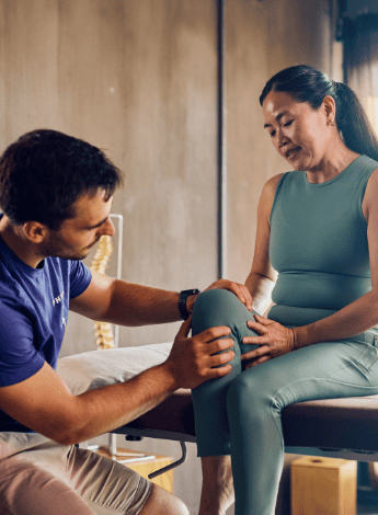 A male physiotherapist is examining the knee of his female patient. She is sitting on an examination table. 